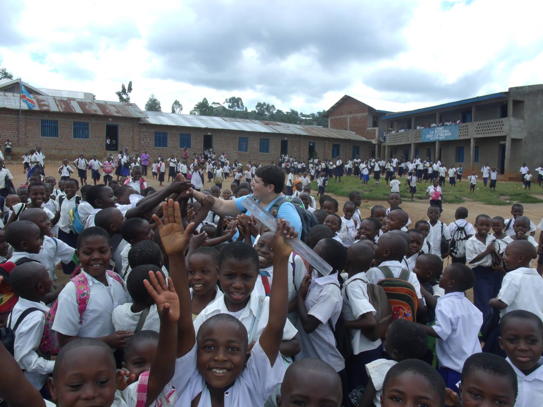 Hundreds of smiling students in white school uniforms crowd around a Kids in the Congo volunteer in a school courtyard, waving at the camera.